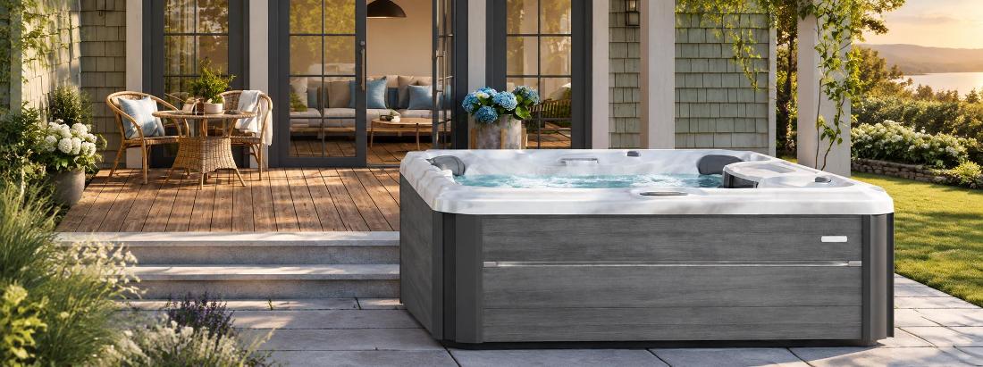 A wide-angle front view of a grey-paneled hot tub on a wooden deck, featuring a white marbled acrylic interior, two grey headrests, and a waterfall feature, set against a backdrop of a modern house with glass doors and a landscaped garden.