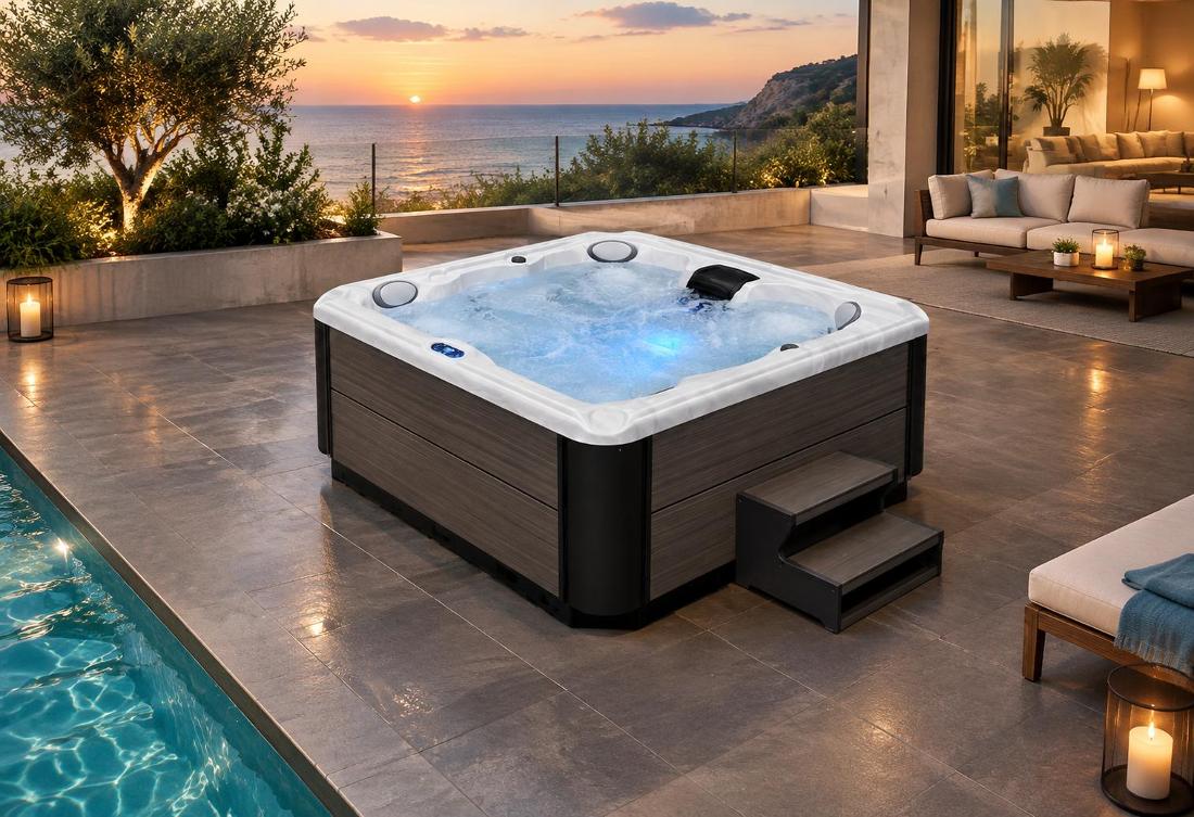 A high-angle 45-degree shot of a square hot tub on a dark stone patio, illuminated by soft blue interior LED lighting, surrounded by modern outdoor furniture and lanterns, with a pool visible in the foreground.