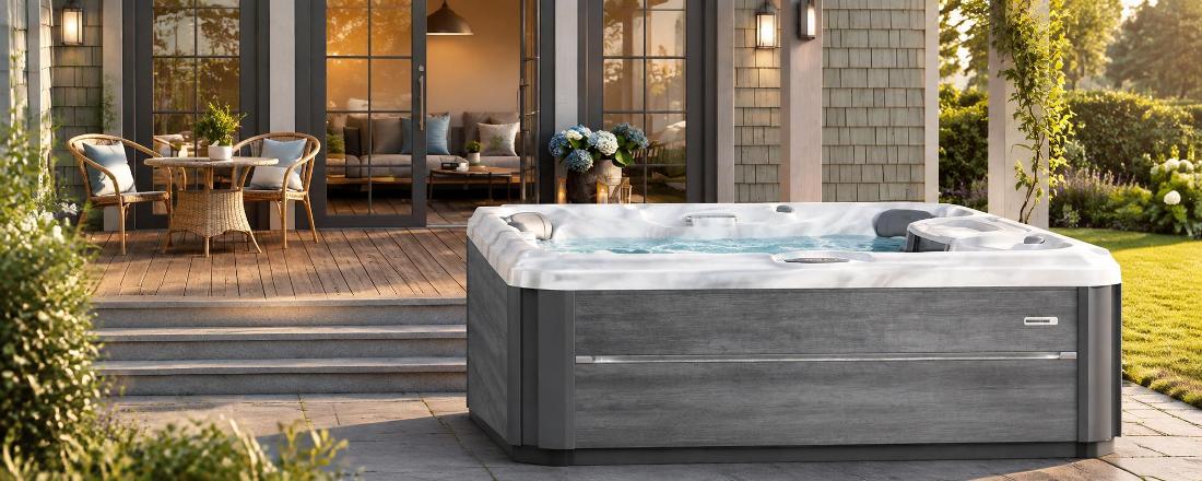 A wide-angle front view of a grey-paneled hot tub on a wooden deck with a white marbled acrylic interior and waterfall feature